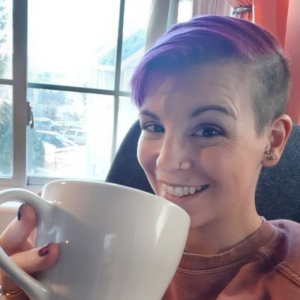 Smiling woman with purple hair and an undercut looks at the camera while drink coffee