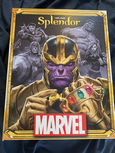 Splendor Marvel. It has a large full colored angry looking Thanos with the Marvel logo located at the bottom in white & red color. There are four Marvel heroes in black & white in the background. They include Dr. Strange, Captain America, Hulk,& Black Widow