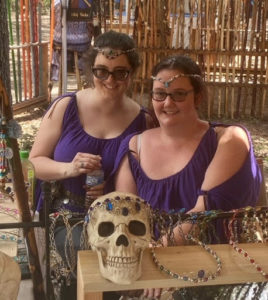 Two women in purple shifts sit behind a skull with a jeweled headpiece. The women are also wearing circlets.