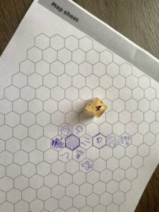 Pictured: A hex sheet from the game with several hexes filled with purple ink representations of the landscapes explored during the game. Also pictured is a six sided Honey & Dice die from Die Hard Dice! 