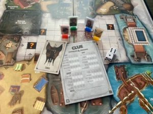 Pictured: Game cards, meeples, dice and Clue sheet are layered over a corder of the game board.