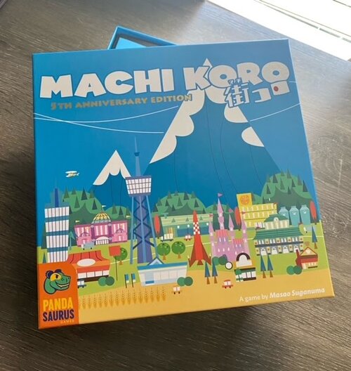 Pictured: The Machi Koro 5th Anniversary Edition game box, featuring a picturesque urban mountain sprawl. There are bright and vibrant buildings against a lush green hilly and blue mountainous backdrop.