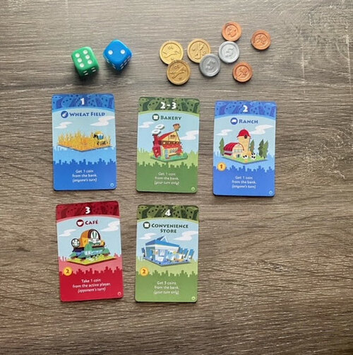 Pictured: Several game cards, including a blue Wheat Field and Ranch card, green Bakery and Convenience Store cards, and a red Cafe card. The two included dice, one green and one blue, and each of the coins, bronze single, silver five, and gold ten piece are also featured.