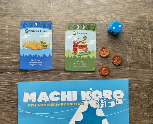 Pictured: The top of the Machi Koro game box, with game cards, coins, and a blue six sided die. The coins are single coin pieces in a bronze color, the cards are the green bakery card, and blue wheat field card.