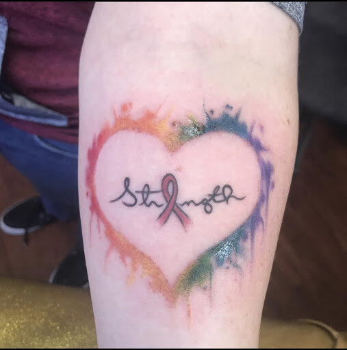 A red, orange, yellow, green, blue, and purple tattoo that symbolizes the rainbow flag with the word “Strength”, the “e” being a pink ribbon in honor of breast cancer awareness.