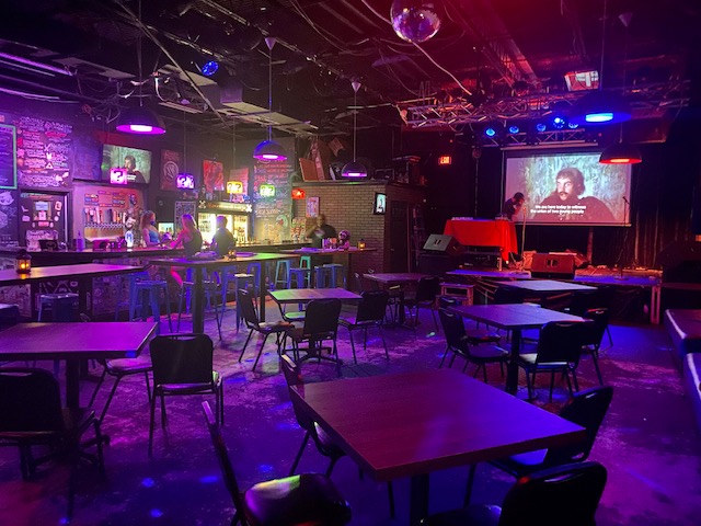 Pictured: The inside of The Geek Easy. Stage and screen in back right, bar on left side. Empty tables, chairs and stools.