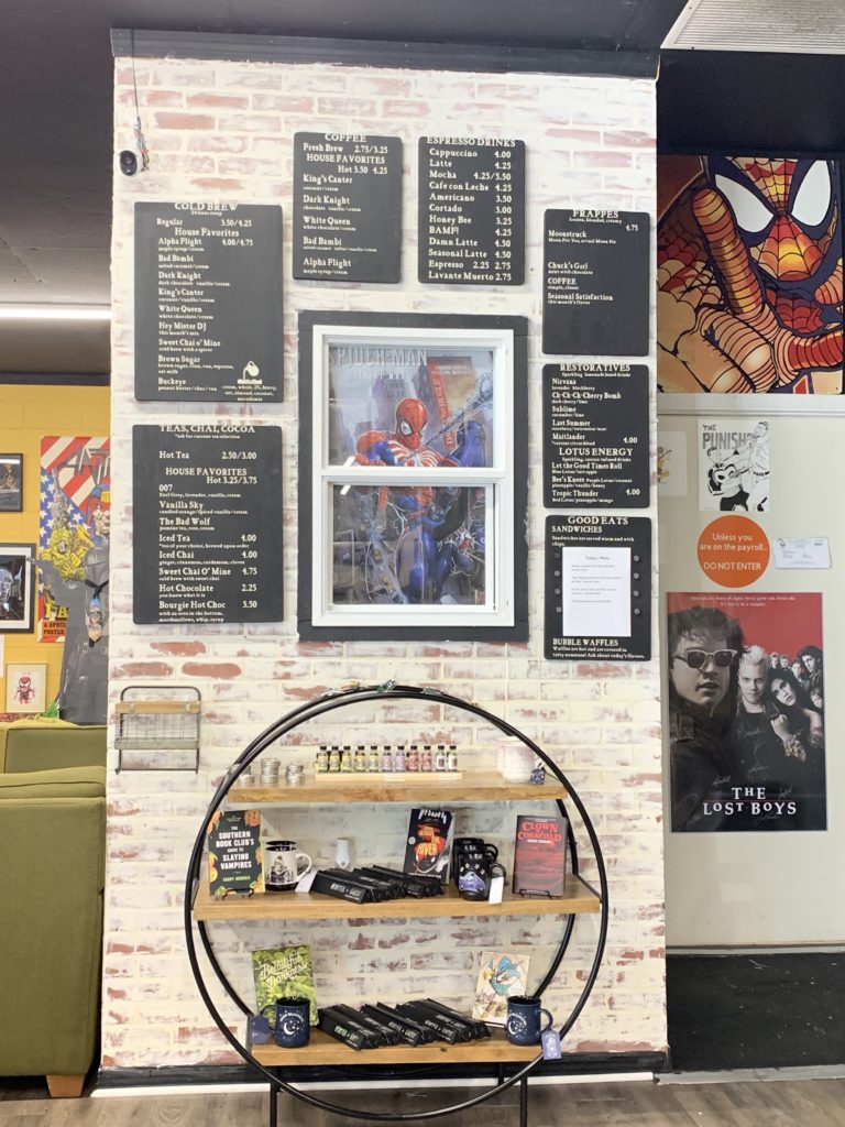 The menu at the BAMF Coffeehouse. A decorative display contains some flavorings and signage, above which hangs a framed Spider-Man poster surrounded by the menu of food and drink offered.