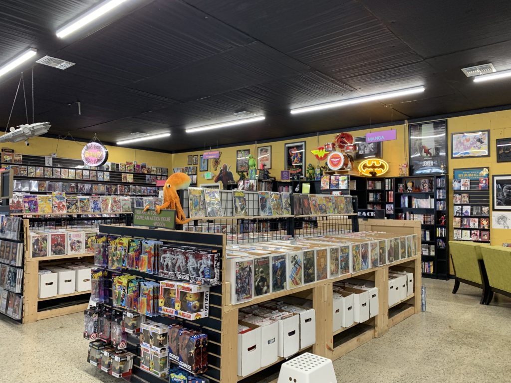 A view of the interior of BAMF Comics & Coffeehouse. The walls are fulled to bursting with comic book heroes and logos on framed posters and neon signs. The shelves are stocked with comics and graphic novels, action figures, stuffed animals and an assortment of collectables.
