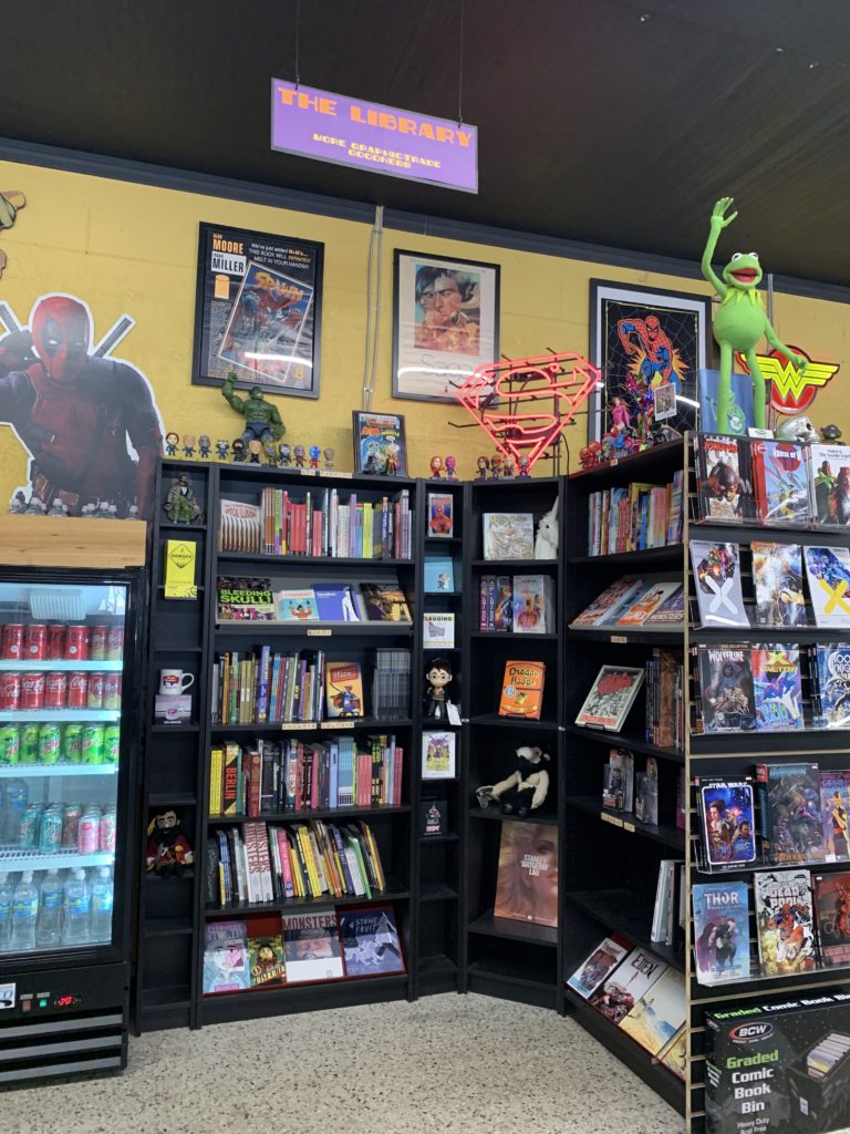 The Library, a portion of BAMF Comics containing books and novels of various genres. Also pictured is a fridge filled with several sodas and fresh water. And of course, a cut out of Deadpool, because no comic shop is complete without a Mere with a Mouth!